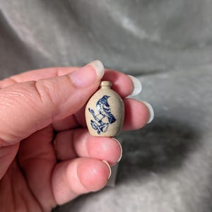 May include: A miniature tan ceramic jug featuring a small blue bird illustration perched on a branch. The jug has a rounded body, a narrow neck, and a small opening at the top. The surface of the ceramic has a speckled texture throughout.