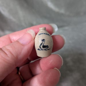 May include: A miniature beige ceramic jug featuring a small, dark blue hand-painted illustration of a swan swimming on water. The jug has a narrow neck and a rounded body, with a faint blue decorative line painted just below the rim.