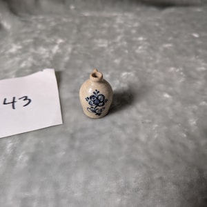 May include: A miniature beige ceramic jug with a narrow neck and a small handle, featuring a hand-painted blue floral design on the front. A white square paper tag with the handwritten number 43 is positioned to the left of the jug.