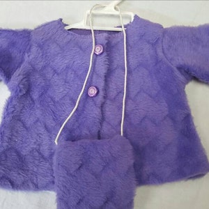 May include: A lavender-colored, plush jacket and matching purse set. The jacket has a quilted pattern and two button closures. The purse has a long, white cord. The set is displayed on a white hanger.