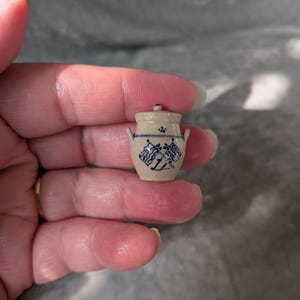 May include: A miniature beige ceramic crock with a lid and two small side handles. The vessel features a blue hand-painted design of two crossed flags on the front, a thin blue horizontal line, and a small blue floral accent near the rim.