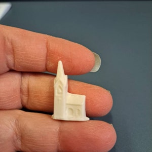 May include: A small, off-white miniature church with a tall steeple and arched windows. The model has a rectangular base and is held between fingers, showcasing its size and detail. The church is a simple, yet elegant representation of a religious building.