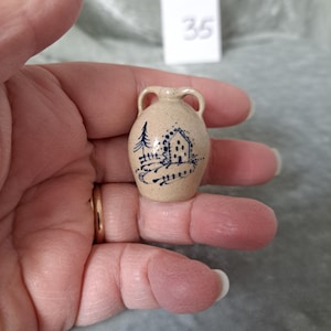 May include: A small, beige ceramic vase with two handles. The vase is decorated with a blue illustration of a house and a tree. The vase is held in a hand.