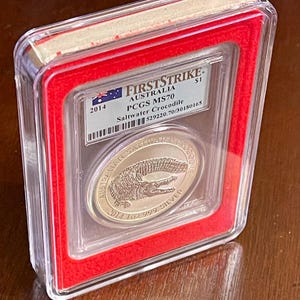 May include: A silver coin encased in a clear plastic holder with a red border. The coin features a crocodile design and the text "AUSTRALIAN SALTWATER CROCODILE". The holder has the text "FIRSTSTRIKE" and "PCGS MS70".