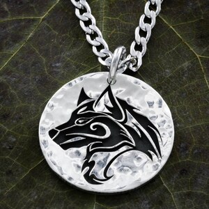 May include: A silver necklace with a round pendant featuring a black wolf head design. The pendant has a hammered texture.