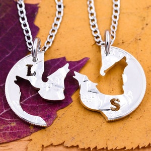 May include: Two silver necklaces with wolf pendants. The pendants are cut out of coins and fit together. The necklaces are on a bed of fall leaves. The pendants are engraved with the letters "L" and "S".
