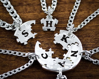 May include: Five silver necklaces with puzzle piece pendants. Each pendant has a different letter, spelling out the word "SHUCKN". The necklaces are on a wooden surface.