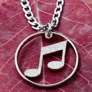 May include: A silver necklace with a music note pendant. The pendant is a cut-out of a music note and is hanging on a silver chain. The necklace is resting on a red leaf.