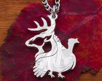 Op de afbeelding: Een zilveren ketting met een hanger met een gedetailleerd silhouet van een kalkoen en een hert. De hanger ligt op een rood blad.