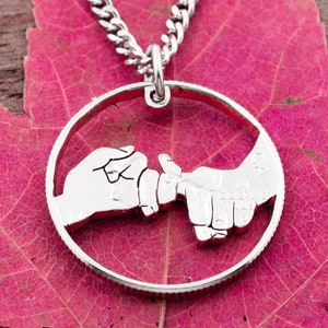 May include: A silver-colored necklace with a pendant featuring two hands interlocked in a pinky promise gesture. The pendant is circular and hangs from a silver chain. The background is a vibrant pink leaf.