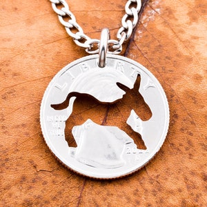 Puede incluir: Collar de plata con un dije de burro recortado. El dije es una moneda de 25 centavos de dólar estadounidense reutilizada con las palabras "In God We Trust" y "Liberty" visibles.