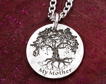 Puede incluir: Un collar de plata con un colgante redondo que presenta un diseño de árbol de la vida con las palabras "Mi Madre" grabadas debajo. El colgante cuelga de una cadena de plata.