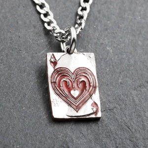 Pode incluir: Colar com pingente prateado com o desenho do Ás de Copas de um baralho. O pingente tem um design de coração vermelho. A corrente é prateada e é exibida em uma superfície escura.