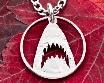 May include: A silver-colored shark tooth pendant necklace. The pendant is a coin cut-out in the shape of a shark's open mouth, with a detailed tooth design. The necklace has a silver chain. The background features red leaves.