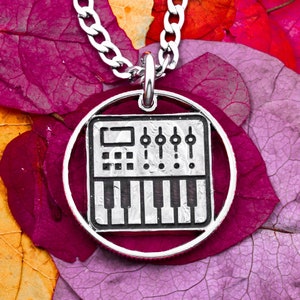 Op de afbeelding: Een zilveren ketting met een ronde hanger met een zwart-witte illustratie van een synthesizer. De hanger is omgeven door een zilveren cirkel.
