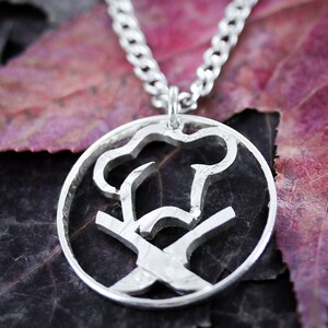 May include: A silver necklace with a round pendant featuring a cutout design of a chef's hat and crossed kitchen knives. The pendant is on a silver chain.