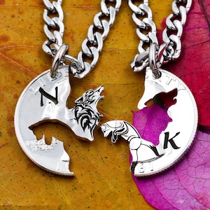 以下が含まれることがあります： 狼の頭のペンダントが付いた銀色のネックレスが2本。ペンダントは半分にカットされており、互いに合わさっています。ネックレスは茶色とピンクの背景の上に置かれています。ペンダントには「N」と「K」の文字が刻まれています。