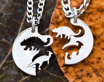 May include: Two silver necklaces with matching pendants. Each pendant is a silver quarter with a dinosaur silhouette cut out. The necklaces are on a brown and green leafy background.