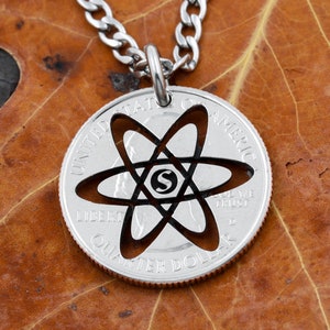 May include: A silver necklace with a silver quarter coin pendant. The coin has a cut-out design of an atom with the letter "S" in the center. The coin reads "United States of America, Liberty, In God We Trust, Quarter Dollar".