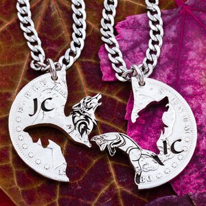 May include: Two silver necklaces with wolf silhouettes cut out of the pendants. The pendants are half of a US half dollar coin with the words "E PLURIBUS UNUM" and "LIBERTY" visible. The necklaces are on a red and brown background.