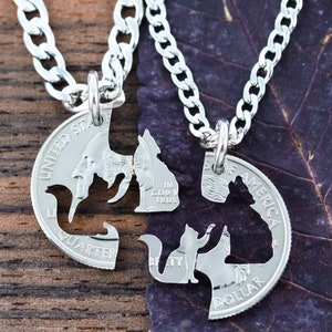 May include: Two silver necklaces with matching pendants. The pendants are cut out of US coins and feature the silhouettes of two cats. One pendant says "United States of America Quarter" and the other says "In God We Trust Liberty Dollar".