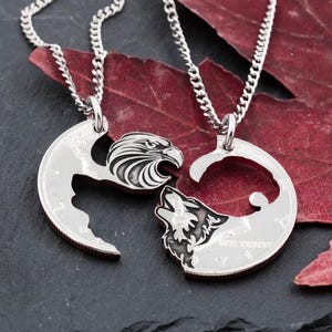 May include: Two silver-toned pendant necklaces. One pendant features an eagle, the other a wolf, each cut from a coin. The pendants are connected to a silver chain. The background includes a dark surface and a red leaf.