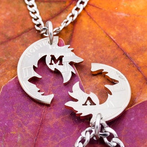 May include: A silver necklace with two pendants shaped like leaves. The leaves are cut out to form the letters "M" and "A". The necklace is on a bed of orange and purple fall leaves.