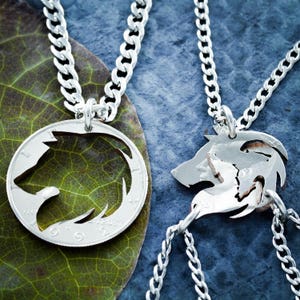 May include: Two silver necklaces with wolf pendant charms. One necklace features a wolf head charm cut out of a silver coin. The other necklace features two wolf head charms connected together.