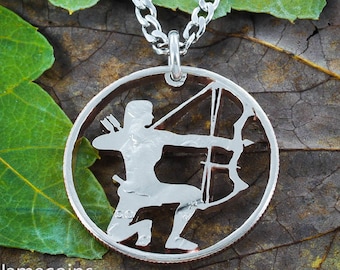 May include: A silver chain necklace with a silver coin pendant featuring a cutout design of an archer with a bow and arrow. The coin is on a brown and green leafy background.