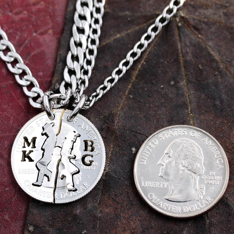 May include: Two silver necklaces with a split quarter coin pendant. The coin has the words "United States of America" and "Quarter Dollar" engraved on it. The coin is split in half, with the initials "MK" on one half and "BG" on the other half.