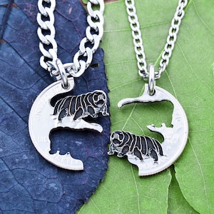 Puede incluir: Dos collares de plata con colgantes a juego. Cada colgante es un semicírculo cortado de una moneda de un cuarto de dólar estadounidense de plata. Un colgante presenta una pequeña criatura negra segmentada, mientras que el otro colgante tiene la criatura recortada, dejando una imagen de espacio negativo.