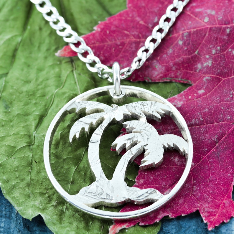 May include: A silver necklace with a circular pendant featuring a cutout design of two palm trees. The pendant is resting on a green and red leaf background.
