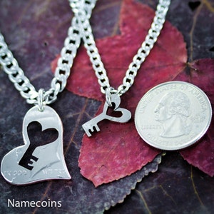 May include: Two silver necklaces, one with a heart pendant with a keyhole cutout and the other with a key pendant with a heart cutout. The necklaces are on a background of red and brown fall leaves.