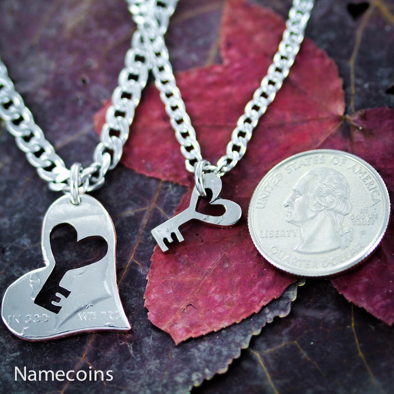 May include: Two silver necklaces, one with a heart pendant with a keyhole cutout and the other with a key pendant with a heart cutout. The necklaces are on a background of red and brown fall leaves.