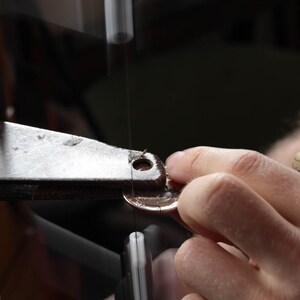 K&ouml;nnte beinhalten: Eine Hand h&auml;lt ein metallisches, m&uuml;nzf&ouml;rmiges Objekt unter einer S&auml;ge. Die S&auml;ge ist an einem Metallrahmen mit einem schwarzen Knopf befestigt.
