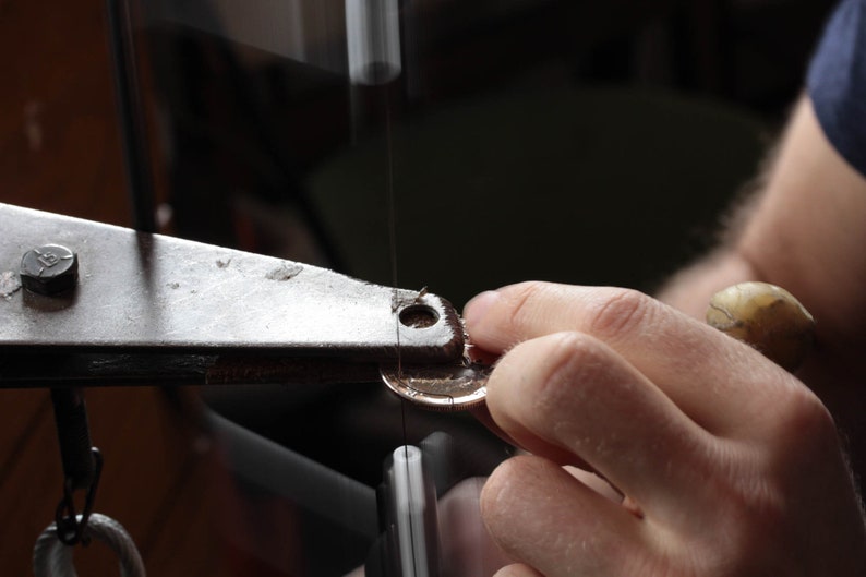 K&ouml;nnte beinhalten: Eine Hand h&auml;lt ein metallisches, m&uuml;nzf&ouml;rmiges Objekt unter einer S&auml;ge. Die S&auml;ge ist an einem Metallrahmen mit einem schwarzen Knopf befestigt.