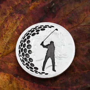 Op de afbeelding: Een zilveren munt met een silhouet van een golfer en een golfbalontwerp. De munt heeft de woorden "In God We Trust" aan de onderkant.