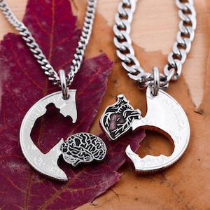 May include: Two silver necklaces with matching pendants. One pendant is a half-dollar coin with a brain cut out, the other is a half-dollar coin with a heart cut out. The pendants fit together.