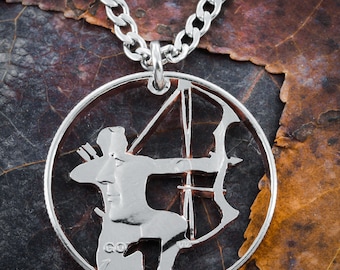 May include: Silver coin necklace featuring a cutout design of an archer with a bow and arrow. The coin is on a brown background.