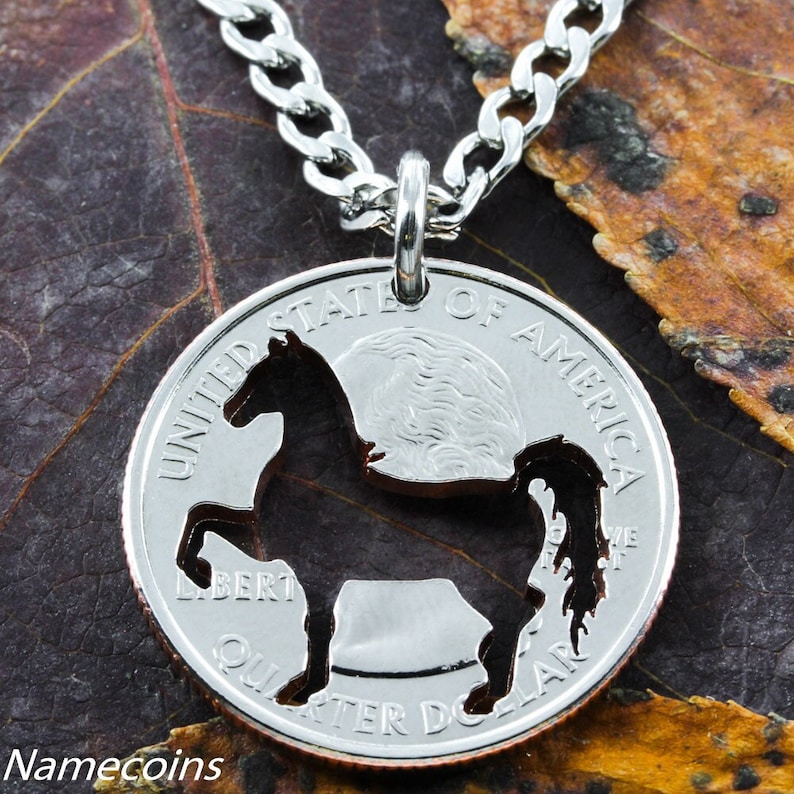 May include: A silver chain necklace with a silver quarter coin pendant. The coin has a horse silhouette cut out of it. The coin is on a brown leaf background.