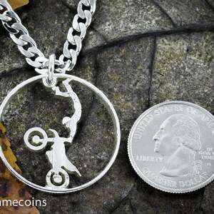 May include: A silver necklace with a silver coin pendant featuring a silhouette of a person performing a stunt on a motorcycle. The coin is attached to a silver curb chain.