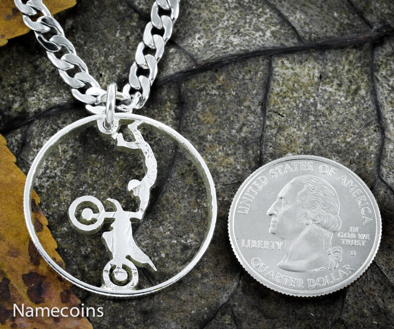 May include: A silver necklace with a silver coin pendant featuring a silhouette of a person performing a stunt on a motorcycle. The coin is attached to a silver curb chain.