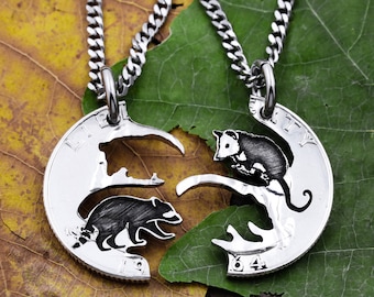 Puede incluir: Dos collares de plata con colgantes a juego que representan un mapache y una zarigüeya. Los colgantes están recortados de una moneda de plata y están conectados por una cadena.