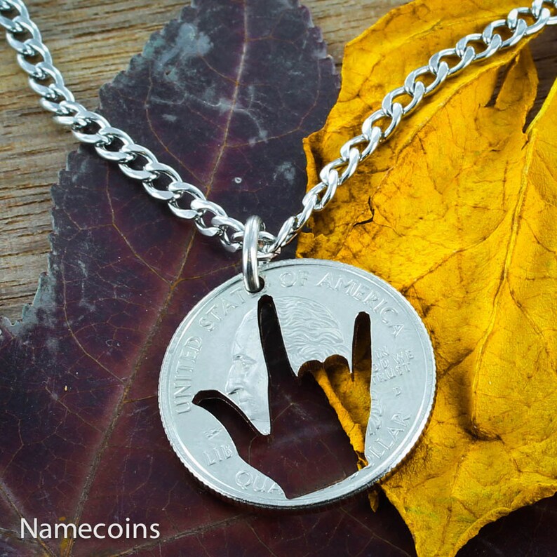 May include: A silver-colored necklace featuring a pendant cut from a quarter coin. The pendant is cut out in the shape of the American Sign Language symbol for "I love you." The necklace is displayed on a leaf.