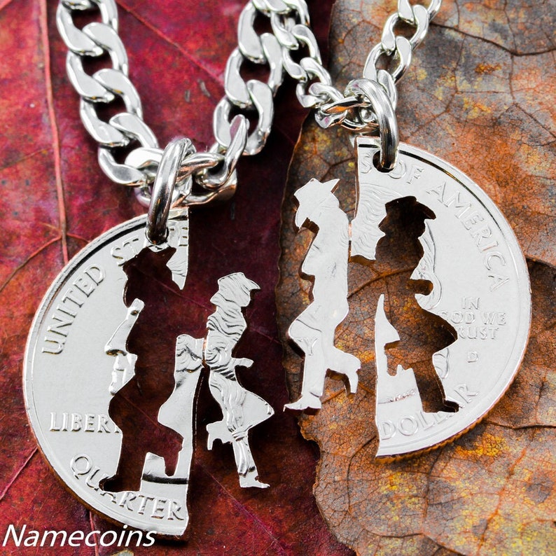 May include: Two silver US quarter dollar coins with a couple dancing silhouette cut out. The coins are attached to silver chains. The coins are on a brown and red leaf.