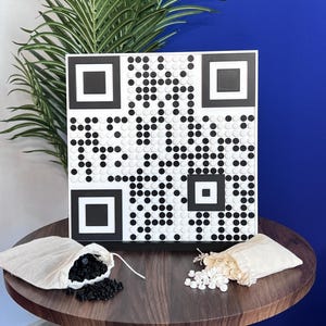 May include: A large, square, black and white QR code art piece made of small, round pieces. Two small canvas bags, one with black pieces and one with white pieces, sit on a dark wood table. A plant and blue wall are in the background.