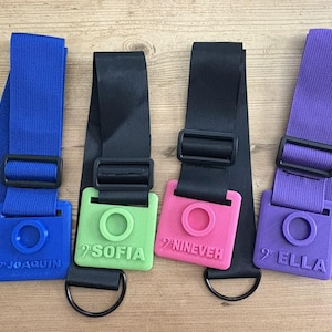 May include: Four adjustable luggage straps with plastic buckles and square tags. The straps are blue, black, black, and purple. The tags are blue, light green, pink, and purple, featuring the names JOAQUIN, SOFIA, NINEVEH, and ELLA embossed on them.