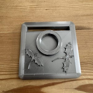 May include: A silver-colored, square 3D-printed plastic stand featuring a central circular ring holder, a rectangular slot at the top, and two embossed dragon figures with spread wings positioned on the left and right sides of the base.