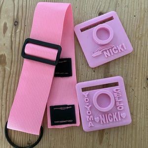 May include: A pink nylon strap with a black plastic buckle and D-ring, accompanied by two square pink plastic attachments. One attachment features a rainbow and the name NICKI, while the other displays the text YO-YOMA CELLO NICKI.