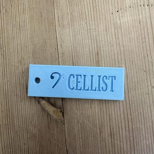 May include: A rectangular light blue tag featuring a circular hole on the left side, a bass clef symbol, and the word CELLIST embossed in capital letters. The surface of the tag displays a textured finish consistent with 3D printed objects.